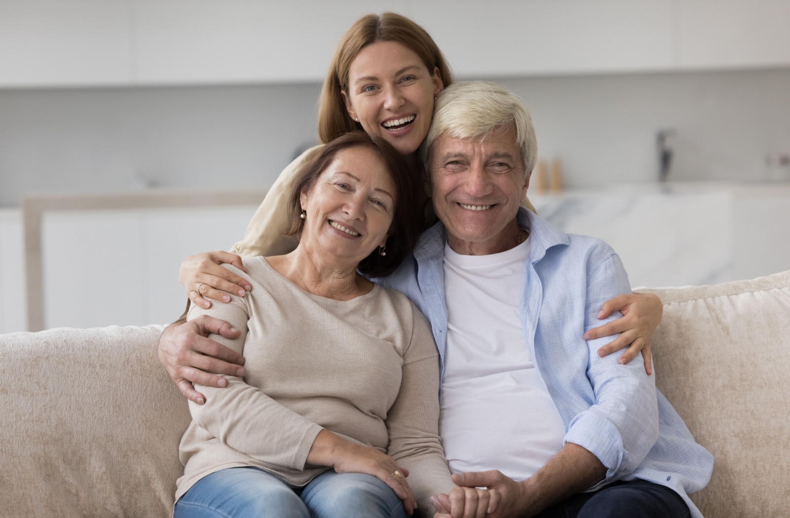 An elderly couple sits on a couch smiling while their adult child stands behind them hugging their shoulders