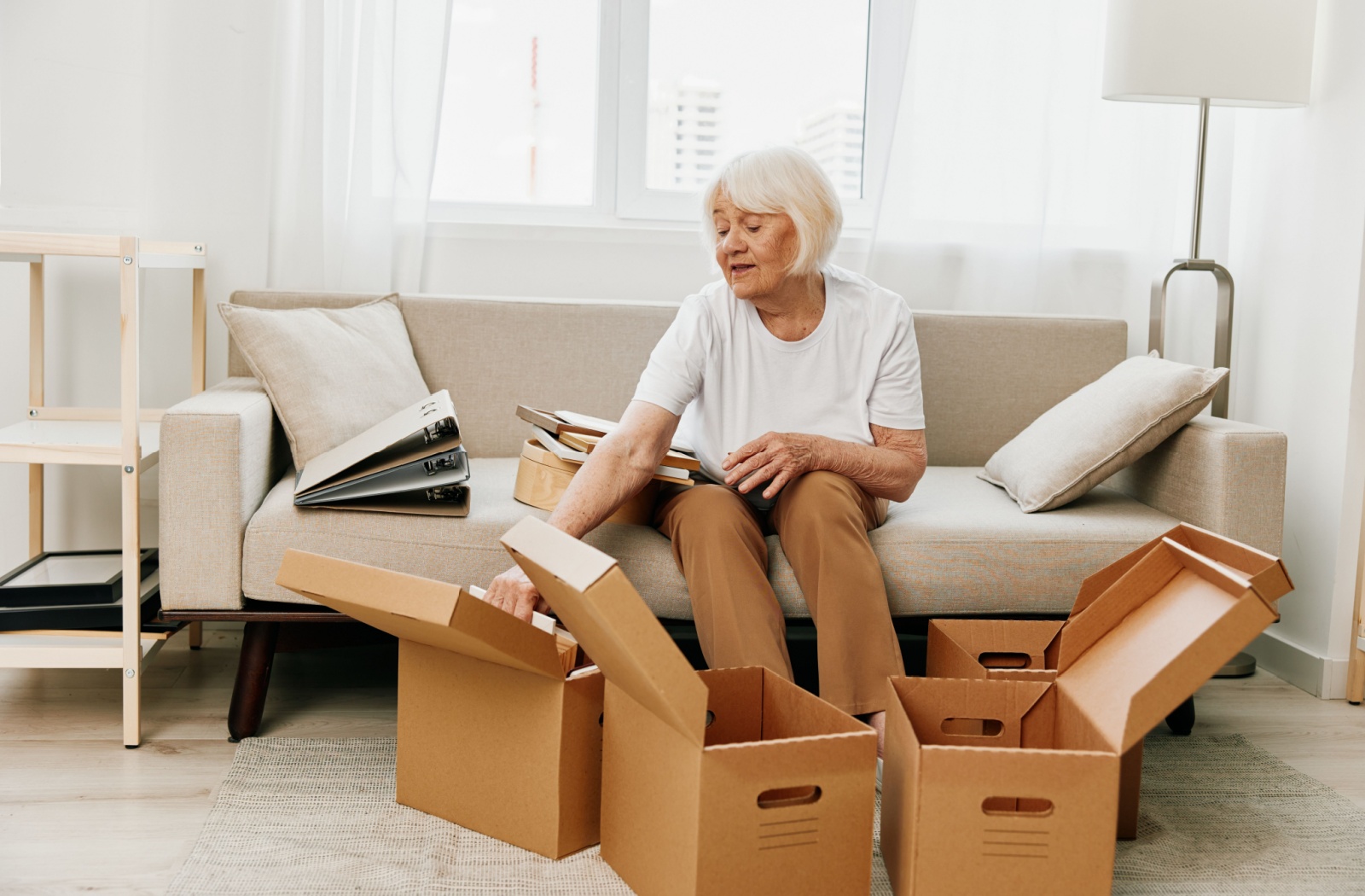 An elderly person sits on a couch and carefully unpacks pictures and photo albums from the four boxes at their feet