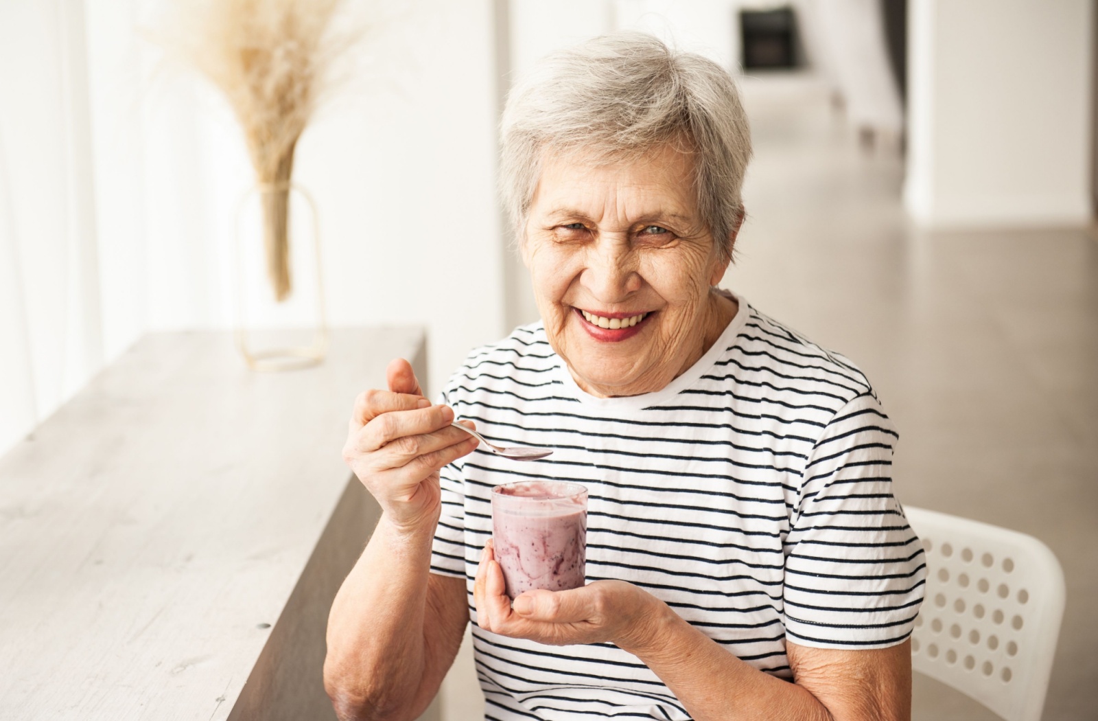 A senior holds a cup of blueberry yogurt with granola, scooping a bite and smiling.