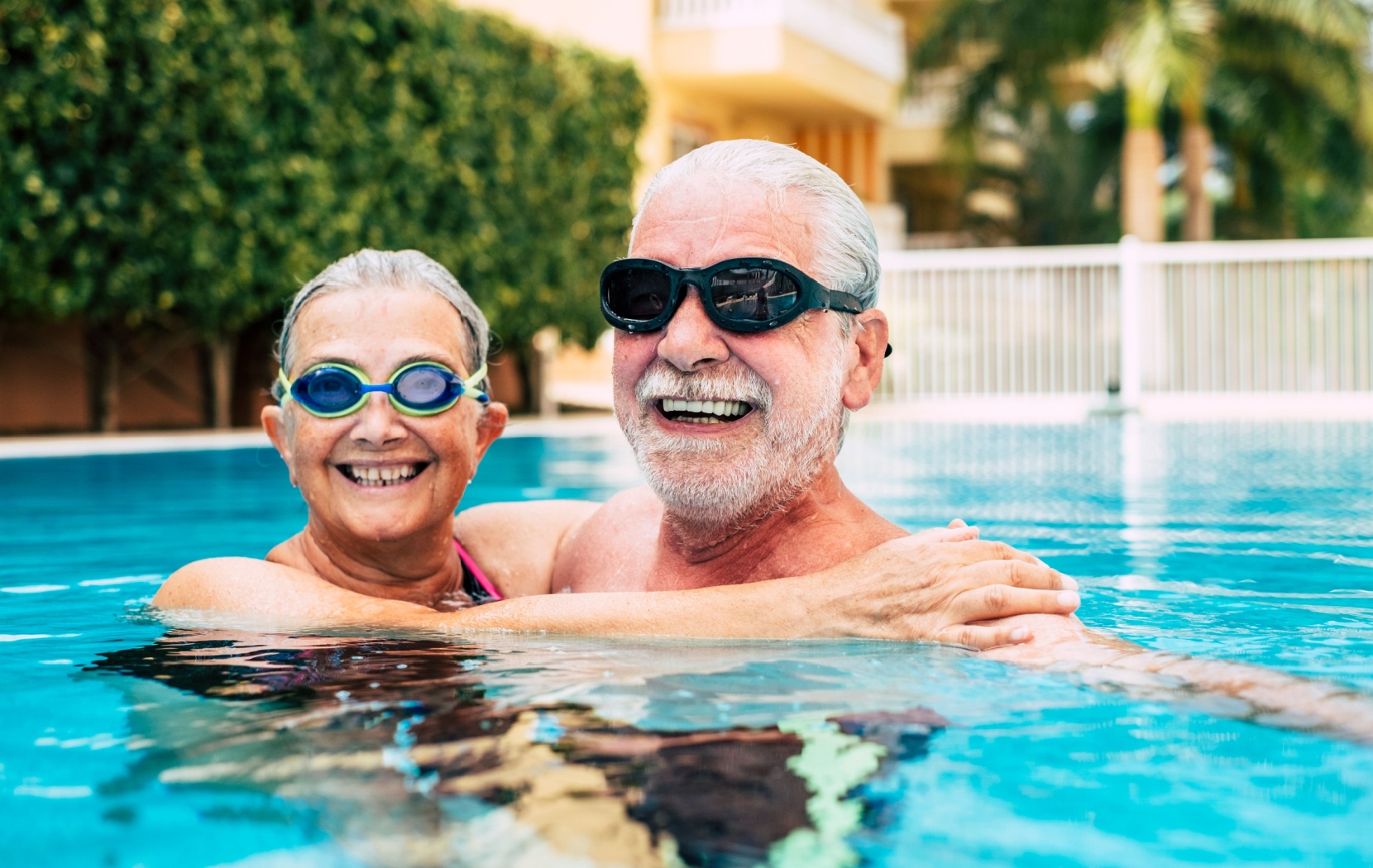 A senior couple wearing swim goggles wrap their arms around each others shoulders as they swim in a pool.
