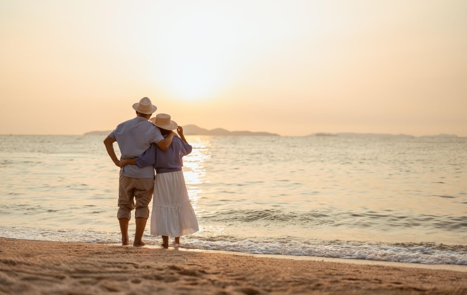 A senior couple stands on a warm beach and stares off into the sunset. Choosing to venture out in the evening to avoid the heat.