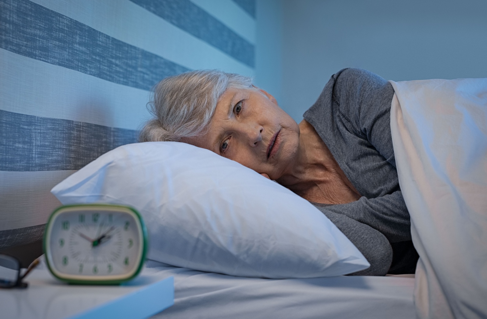 A frustrated older adult staring at their alarm clock late at night while lying awake in bed.