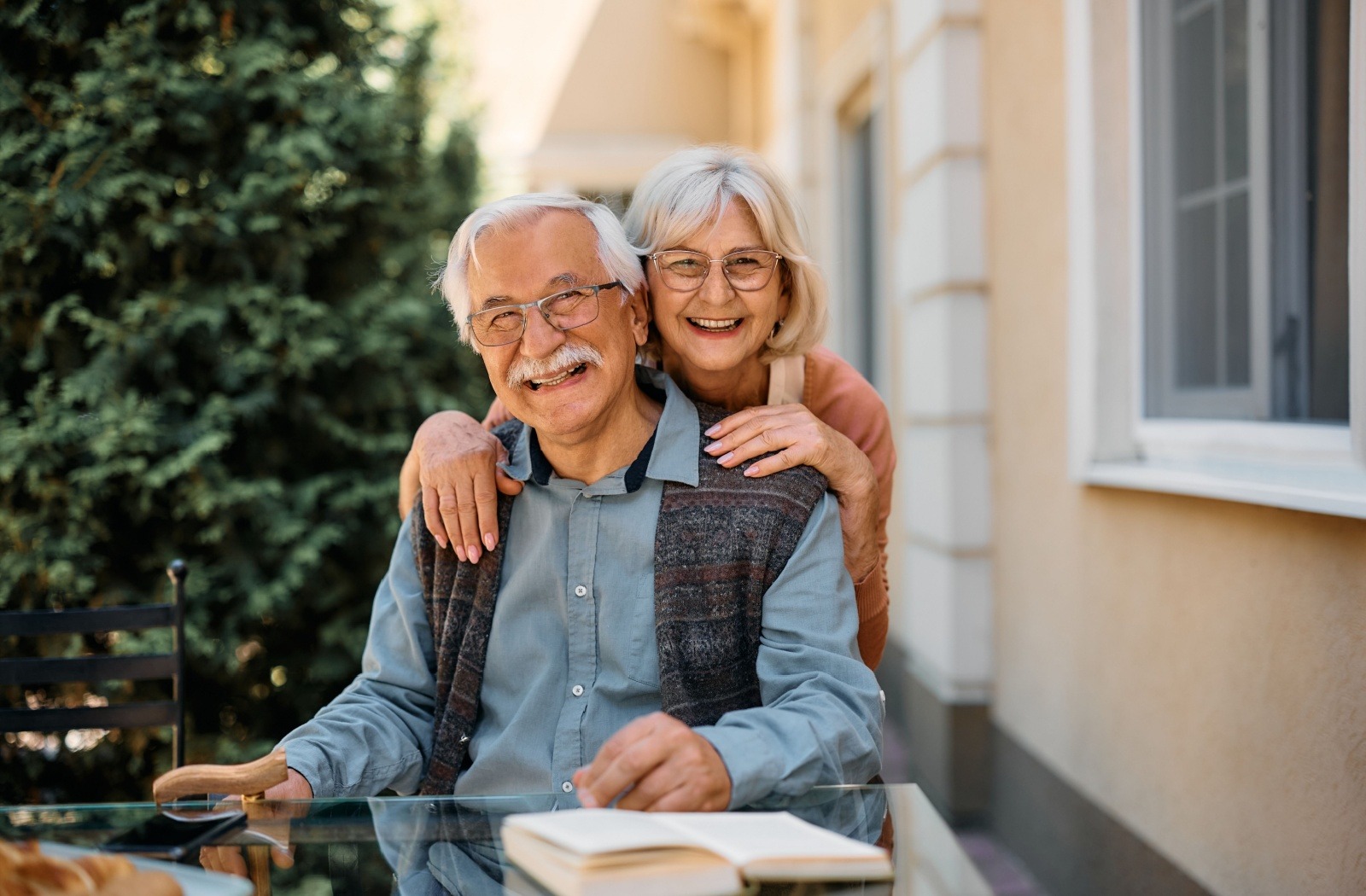 A senior sits at an outdoor table and smiles at the camera as their spouse embraces them from behind